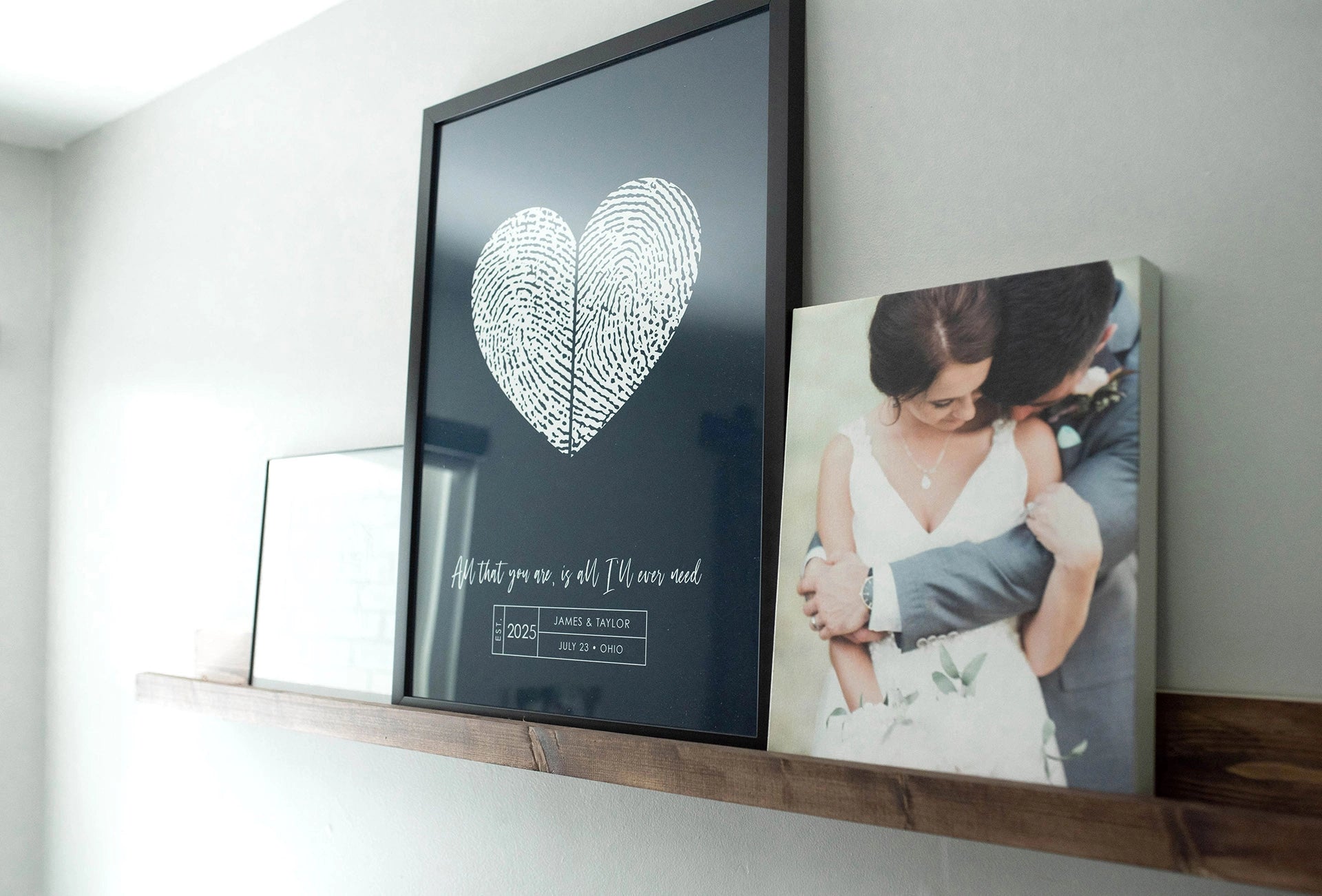 Black-framed print with a white fingerprint heart and sentimental text sits on a wooden shelf, beside a wedding photo, conveying love and intimacy.