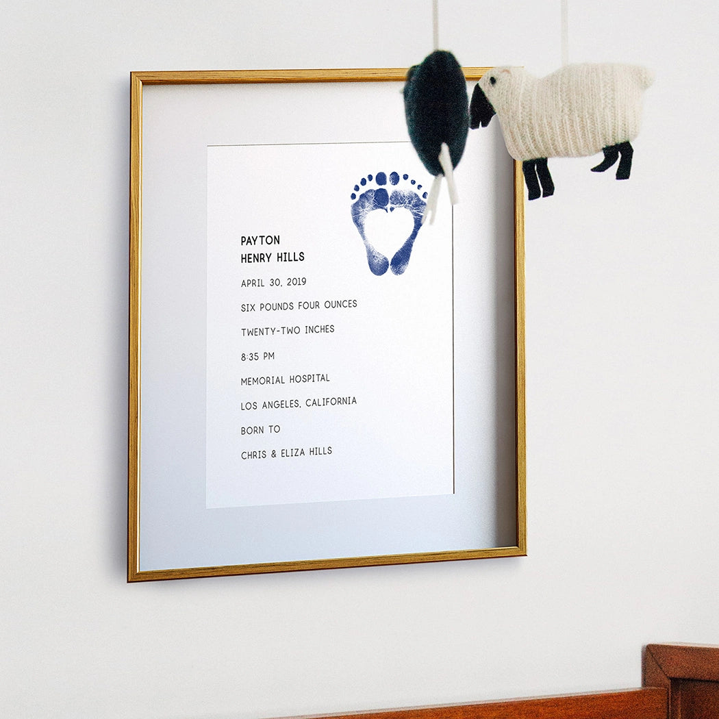 Framed birth stats announcement with blue baby footprints and details, above a wooden crib. A soft lamb mobile hangs, creating a warm, nurturing atmosphere.