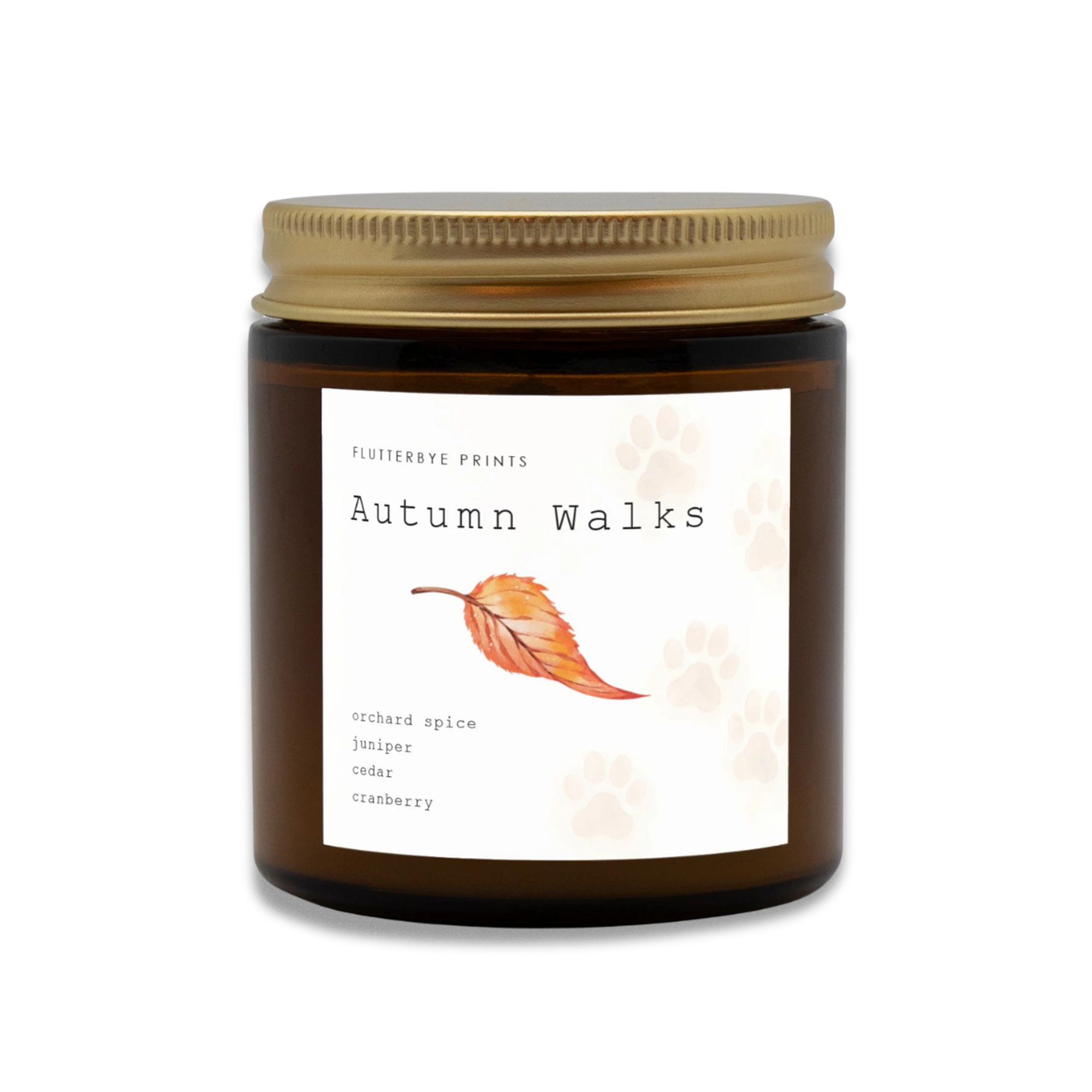 Amber candle jar with a gold lid labeled "Autumn Walks" features a red fall leaf design and faded watercolor dogs paw print. Scent notes: orchard spice, juniper, cedar, cranberry. Cozy and warm.