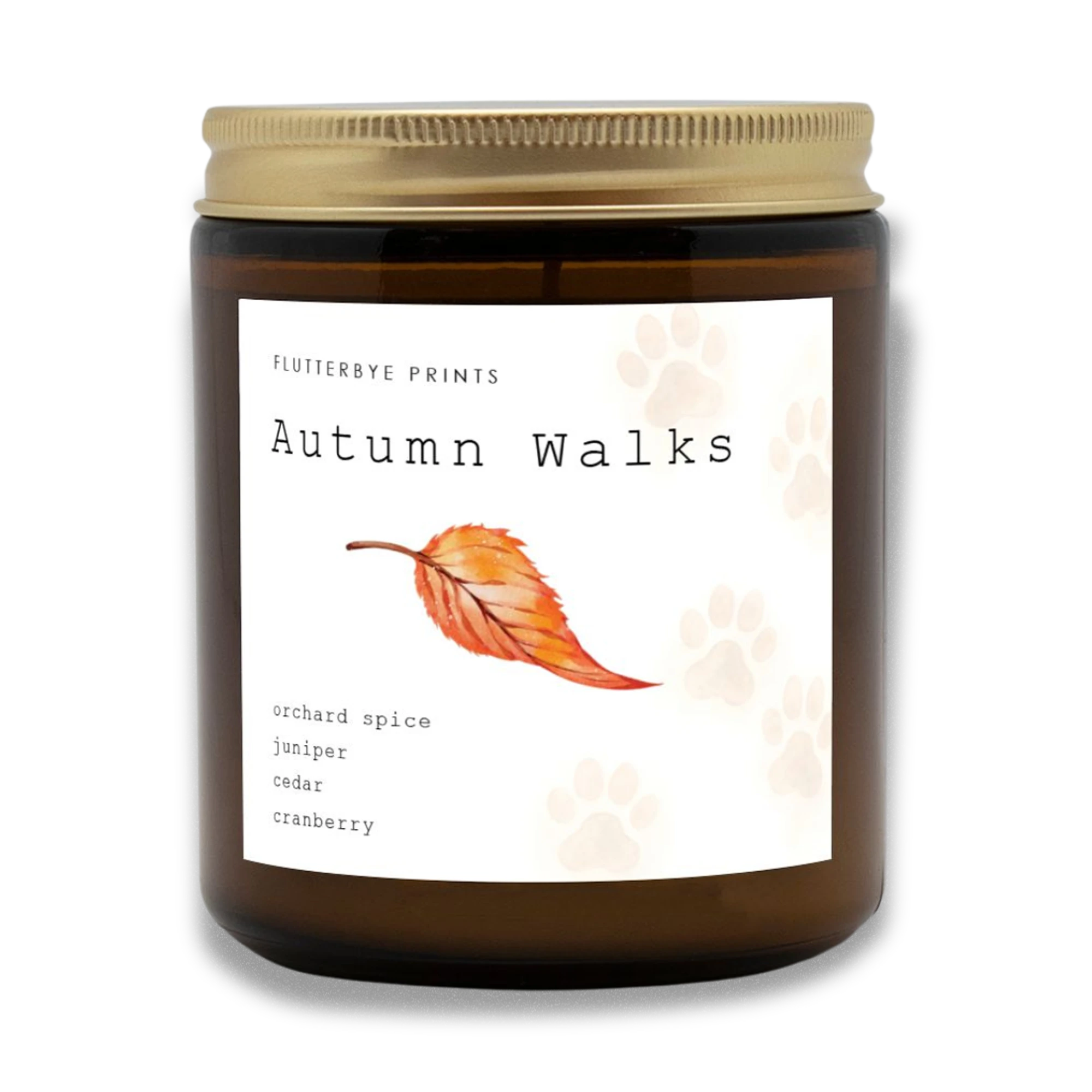 Amber jar candle labeled "Autumn Walks" with a golden lid. The label features an orange leaf, dog paw prints, and scents: orchard spice, juniper, cedar, cranberry.
