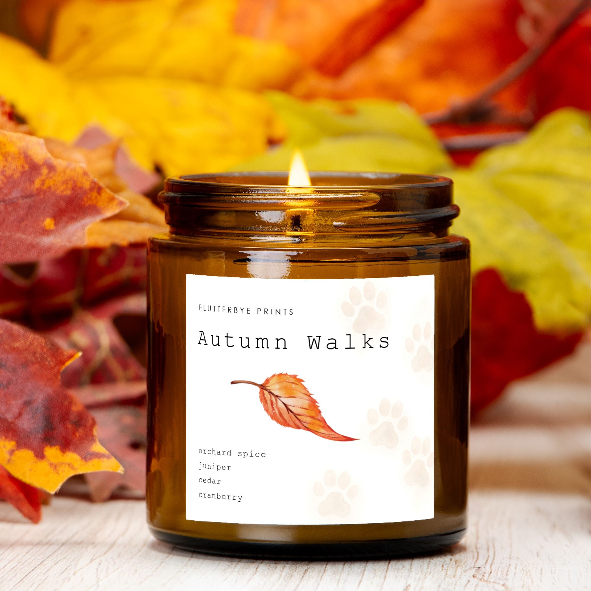 A lit candle labeled "Autumn Walks" in a brown jar, surrounded by vivid yellow and red fall leaves. The label features a leaf, dog paw prints and lists scents like orchard spice and cedar. Cozy and warm tones.