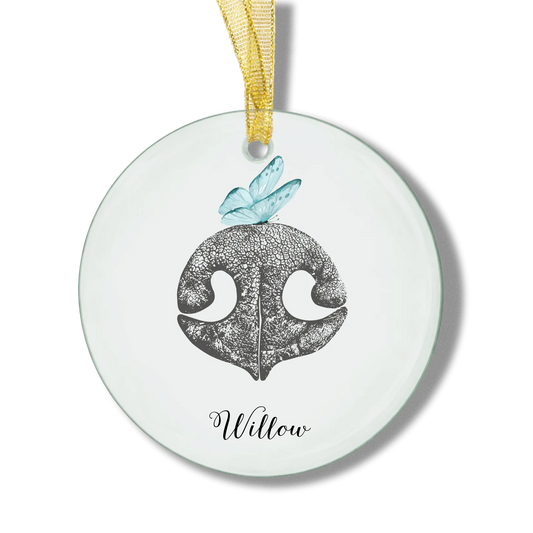 A round glass ornament with a gold ribbon features an illustration of a textured, gray dog nose print and a blue butterfly. The pet name "Willlow" is inscribed below.