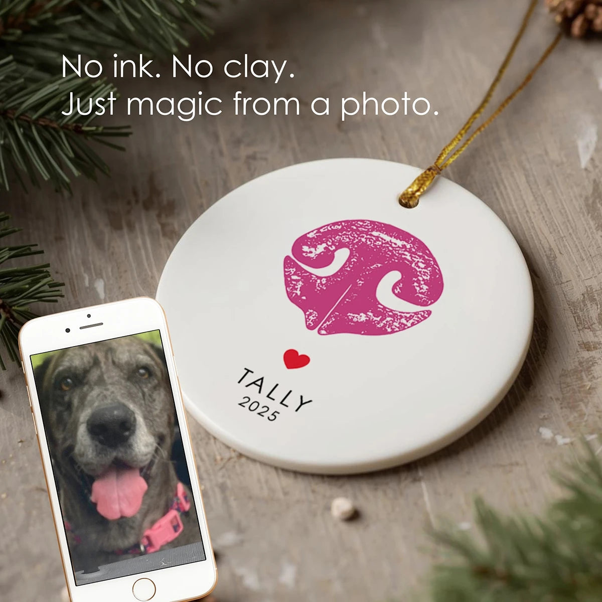 A round ornament with a pink dog nose print and the name “Tally 2025” lies on a wooden surface. A phone displaying a dog’s nose is nearby. Spruce branches add a festive touch. Text reads, "No ink. No clay. Just magic from a photo."