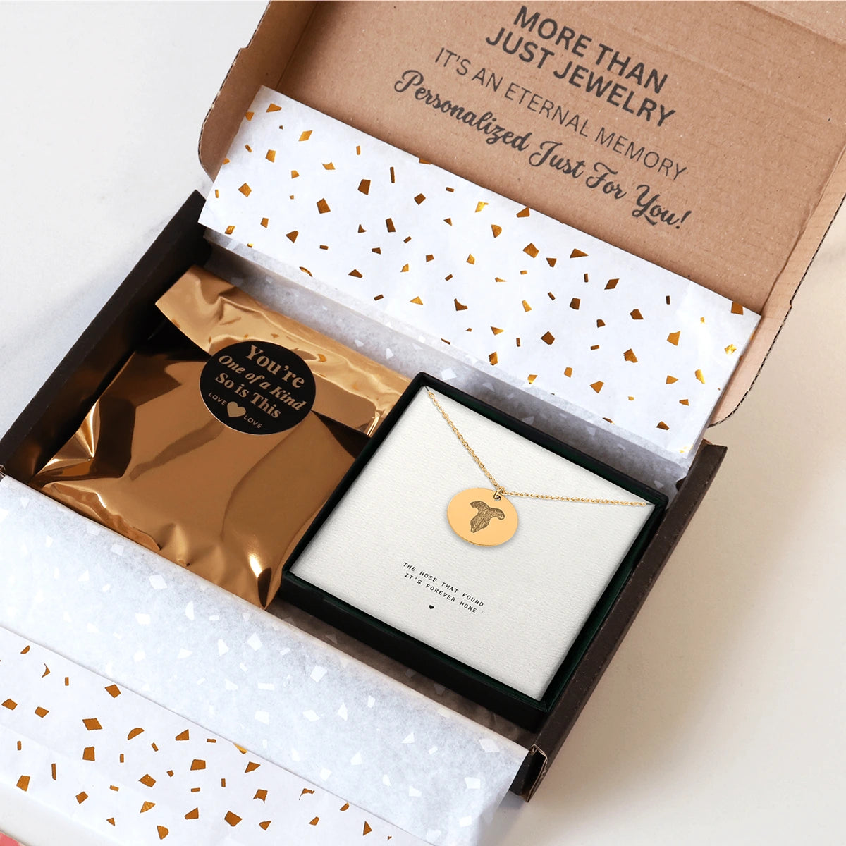 An open jewelry box lined with gold and white confetti paper contains a gold pendant cat nose print memorial necklace and a shimmering gold package. The box lid reads, "More than just jewelry. It's an eternal memory personalized just for you!" The scene conveys a sense of luxury and personalization.
