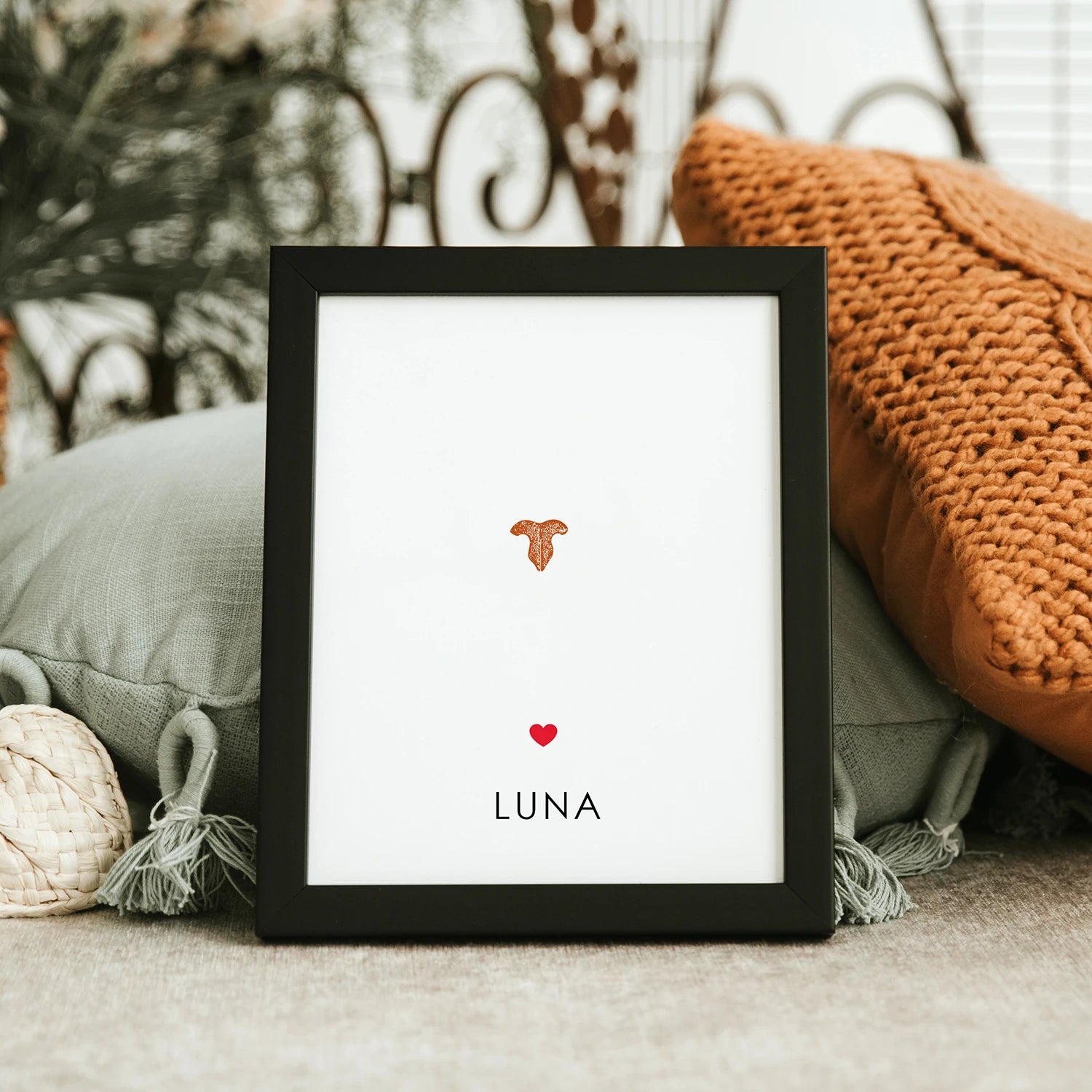 Framed minimalist pet memorial gift art with cat's nose print in orange and the name "Luna" below a red heart. Background includes cozy pillows.