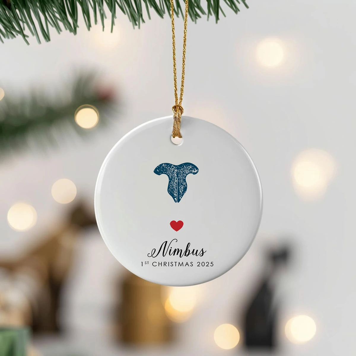A white circular ornament hangs from a Christmas tree branch. It features a blue cat nose print, a red heart, and the text "Nimbus 1st Christmas 2025" below. The background is softly blurred with warm lights, conveying a festive and cozy atmosphere.