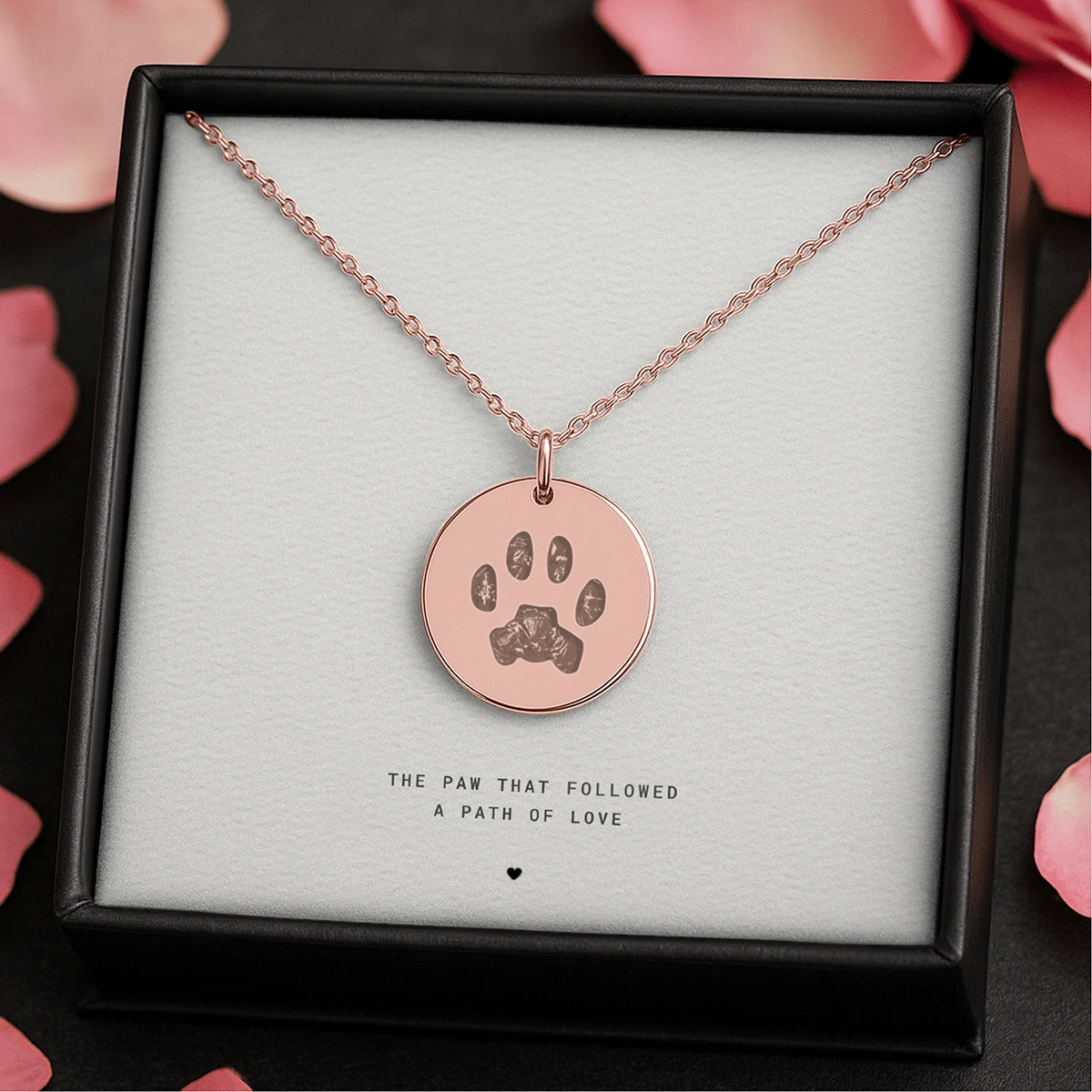 Rose gold necklace with a pendant featuring a cats paw print, set in a gift box with the phrase "The paw that followed a path of love" and surrounded by pink petals.