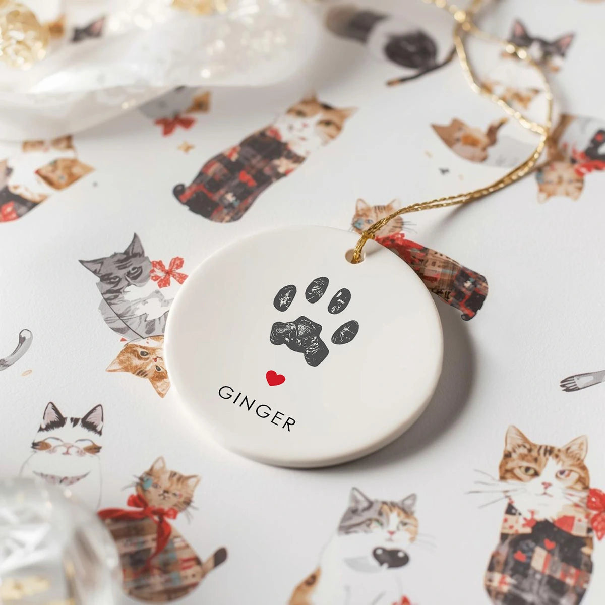 Round holiday pet ornament with a cat paw print and the name "Ginger" below a red heart. It rests on paper printed with illustrations of cats wearing festive outfits.