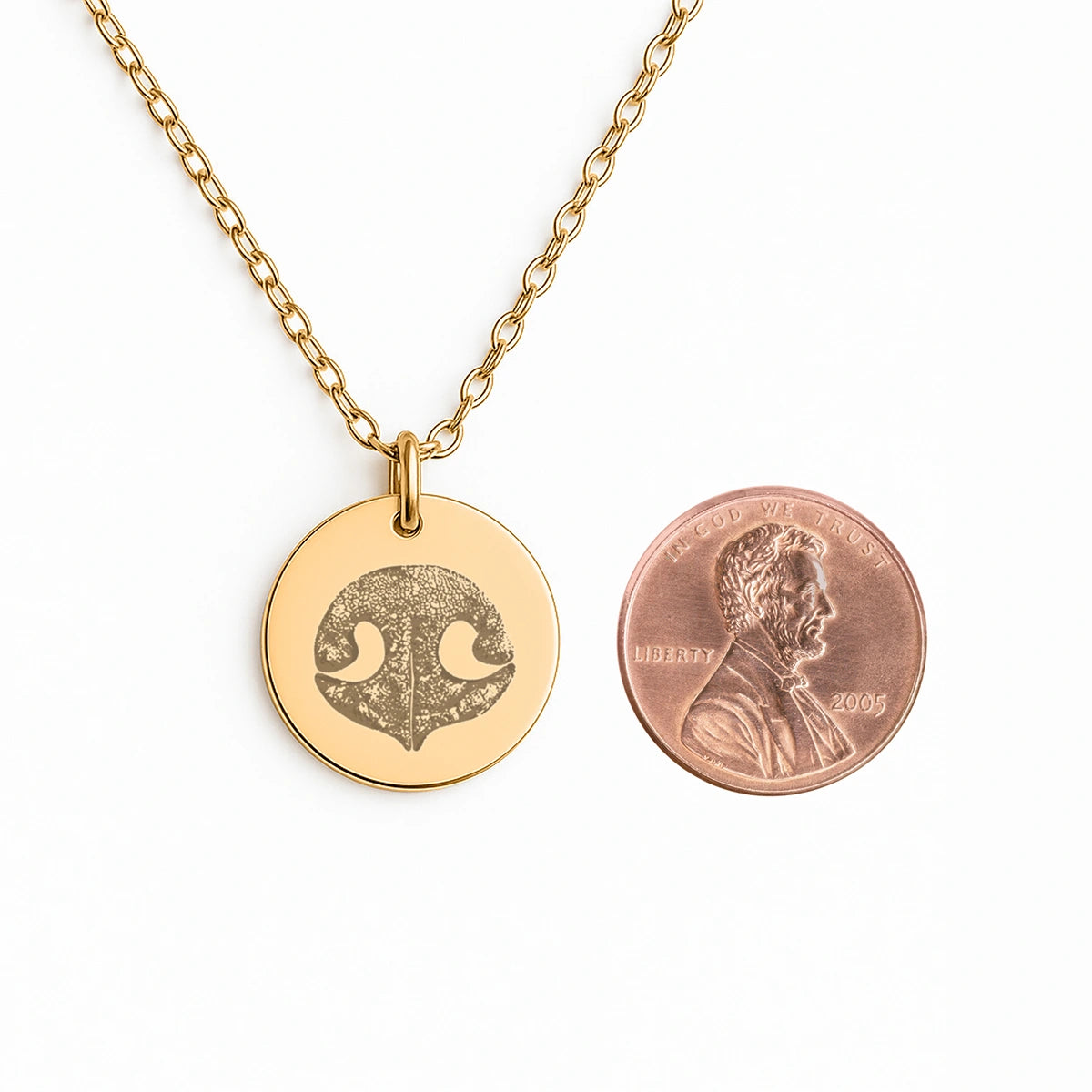 Gold necklace with a round pendant featuring a dog nose print design beside a penny for size comparison. Elegant and minimalist pet memorial jewelry.