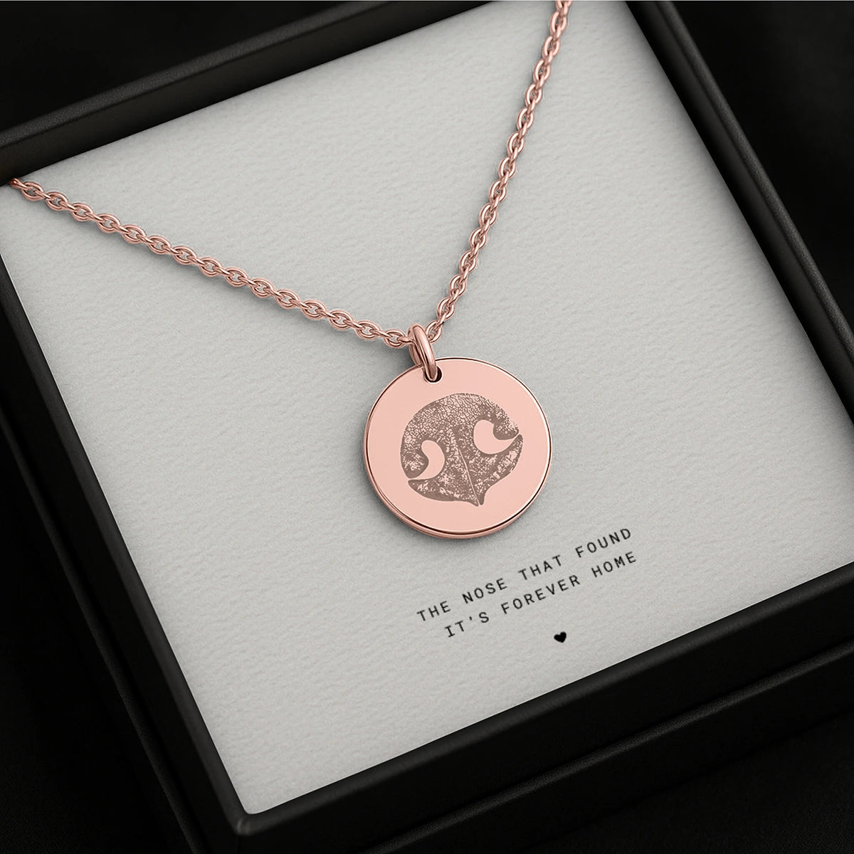 Rose gold necklace with a round pendant featuring a dog's nose print, set against a white background. The text reads, "The nose that found its forever home."