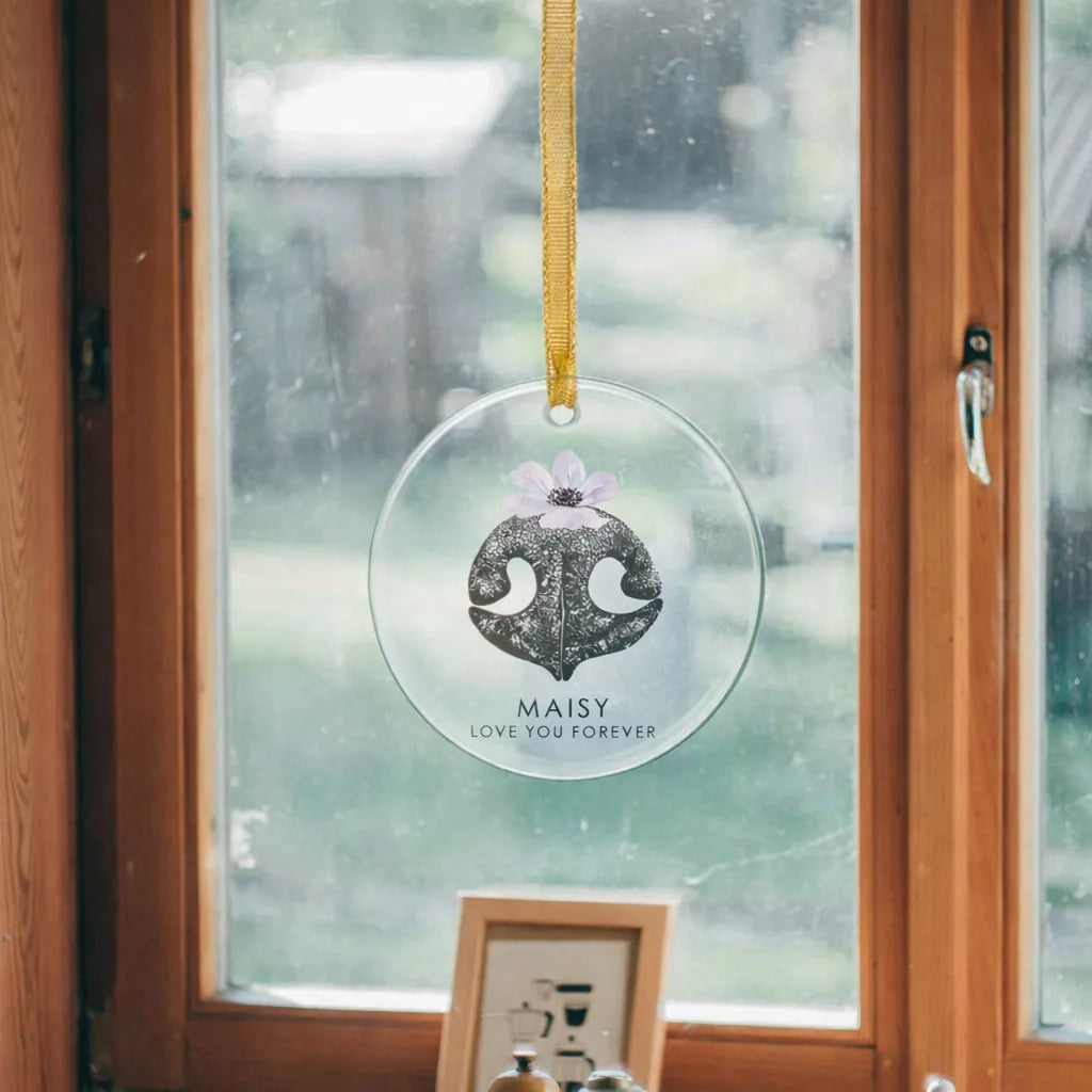 Clear glass ornament with a dog nose print and purple flower hangs by a gold ribbon in front of a window. The words read "Maisy, Love You Forever."