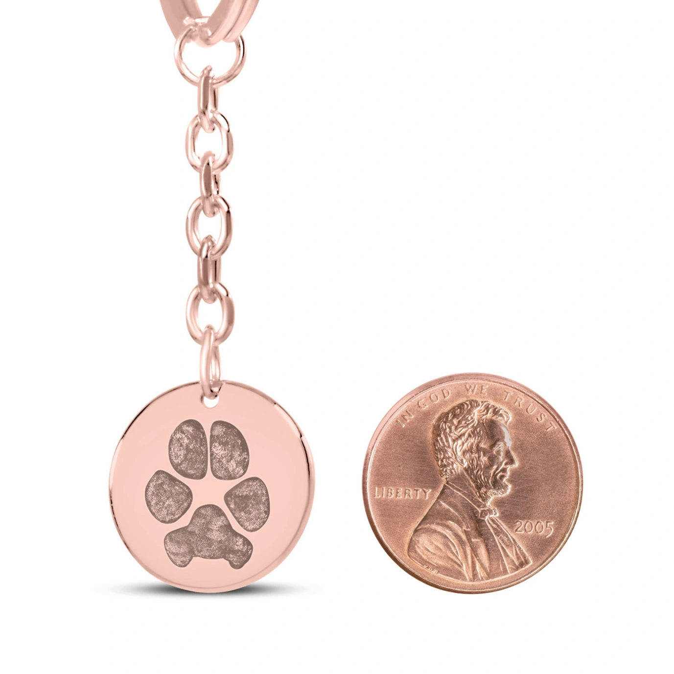 Rose gold coin pendant keychain with featuring an engraved dog paw print design, displayed next to a penny for size comparison.