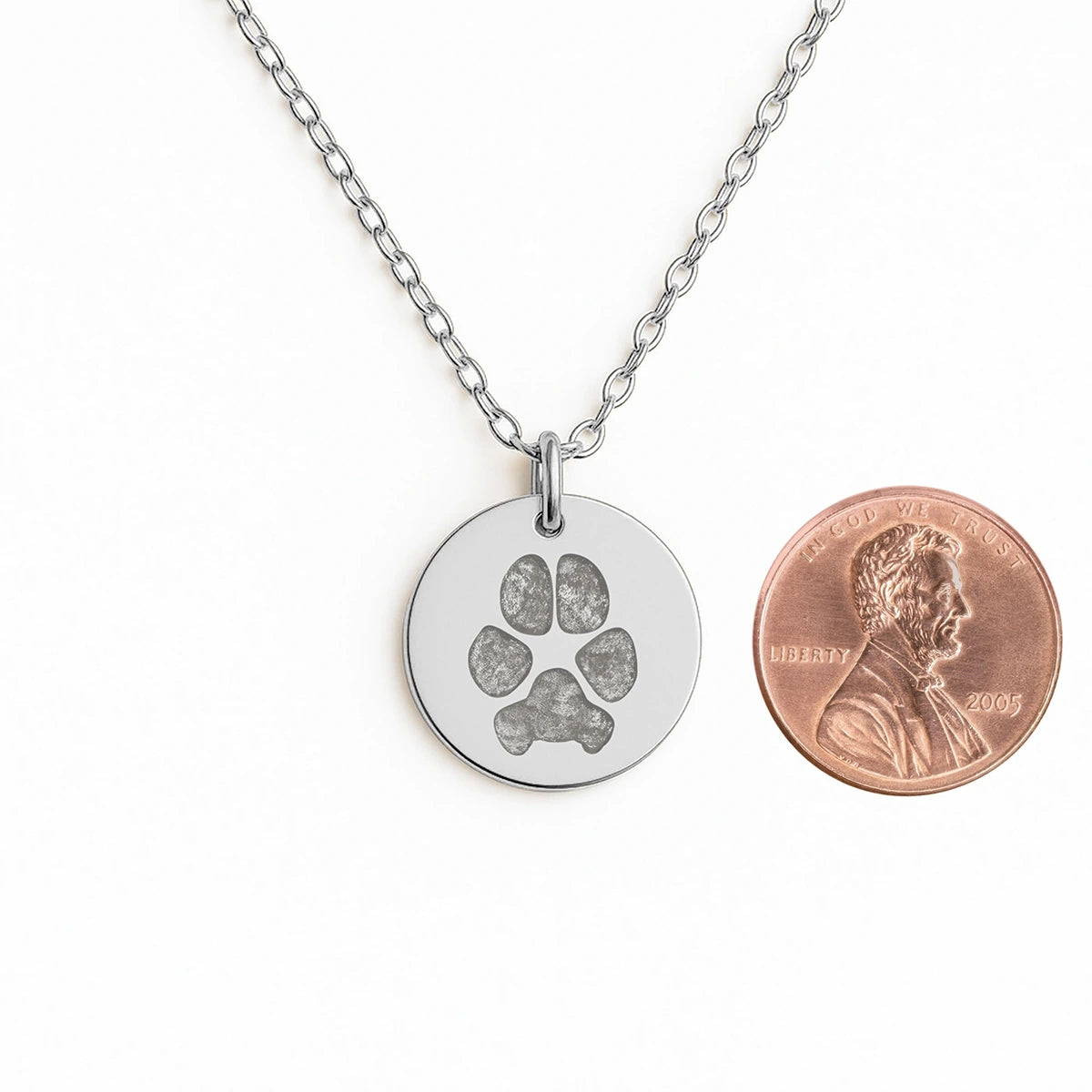 Silver coin pendant necklace with featuring an engraved dog paw print design, displayed next to a penny for size comparison. Elegant and simple.