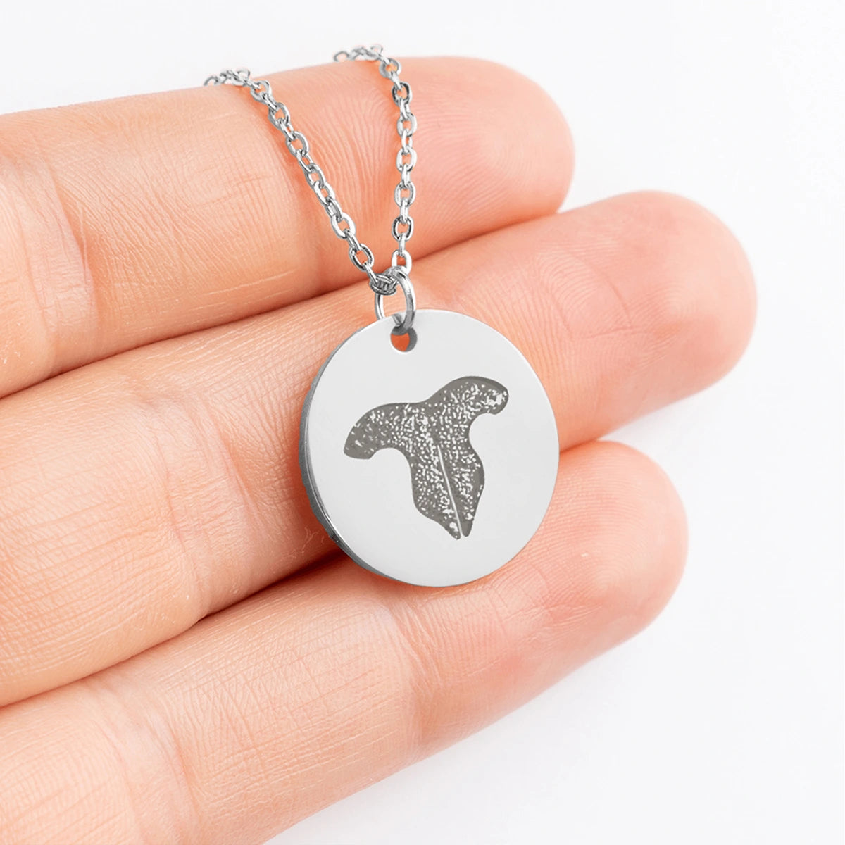 A hand holds a silver chain necklace with a round pendant featuring an engraved cat nose print design. The background is white, highlighting the pendant.