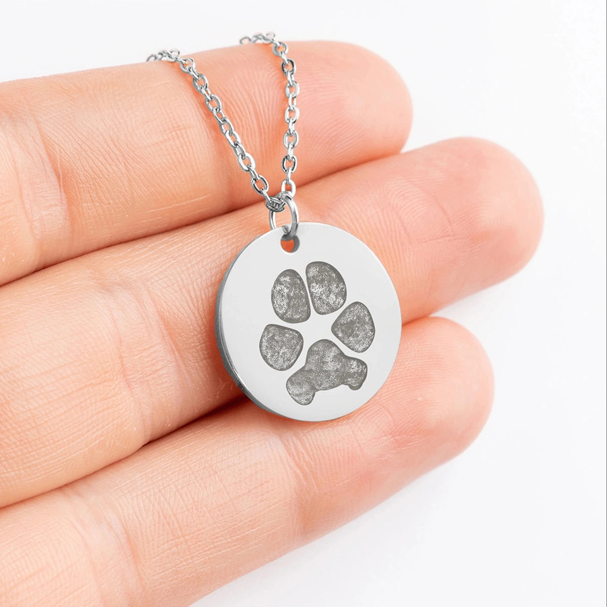 A hand holding a silver necklace pendant with a dog paws printing engraving. The pendant is circular and shiny, conveying a sense of pet love and elegance.
