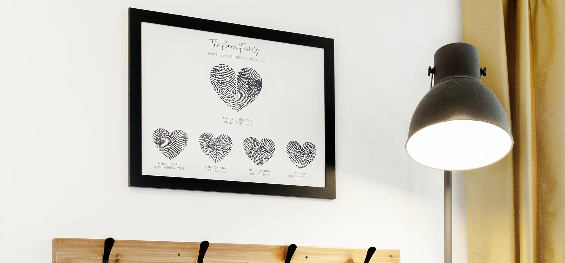 Framed fingerprint heart artwork with family names and dates, beside a modern floor lamp and wooden coat rack with a yellow jacket hanging. Cozy atmosphere.