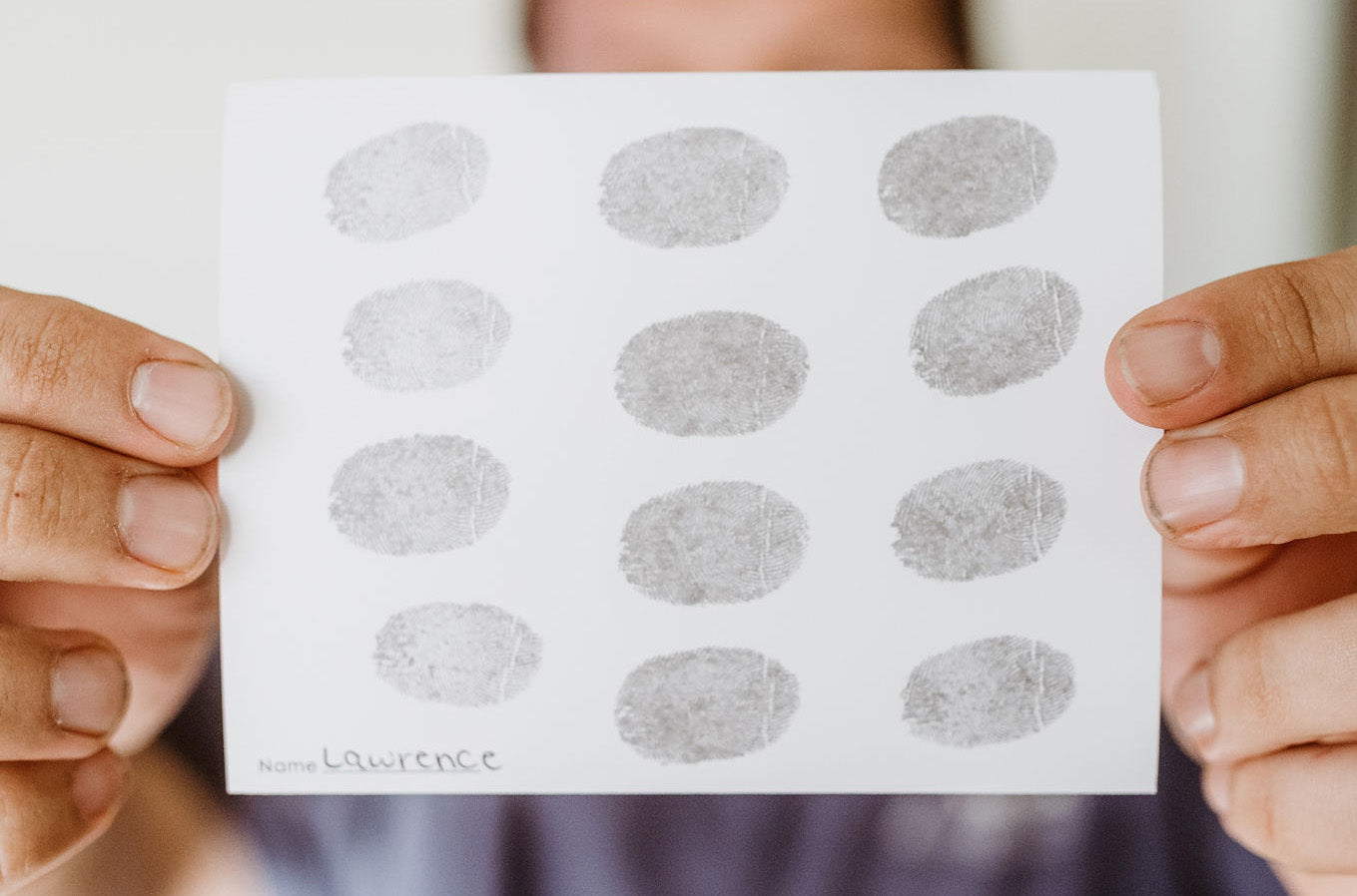 A person holds a sheet with twelve fingerprints in rows. The name 