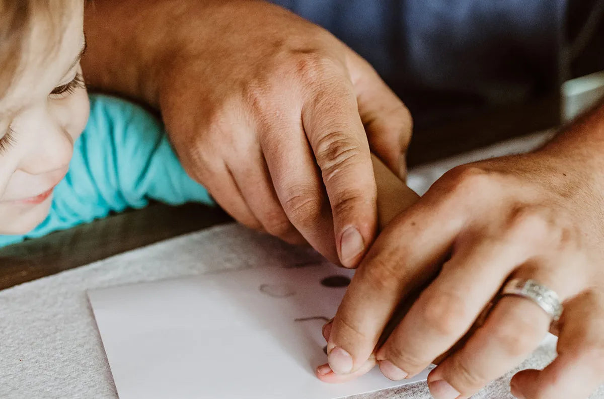 Close-up of a parent’s hands guiding a child’s fingers to draw on paper. The child smiles, wearing a blue shirt, conveying warmth and bonding.