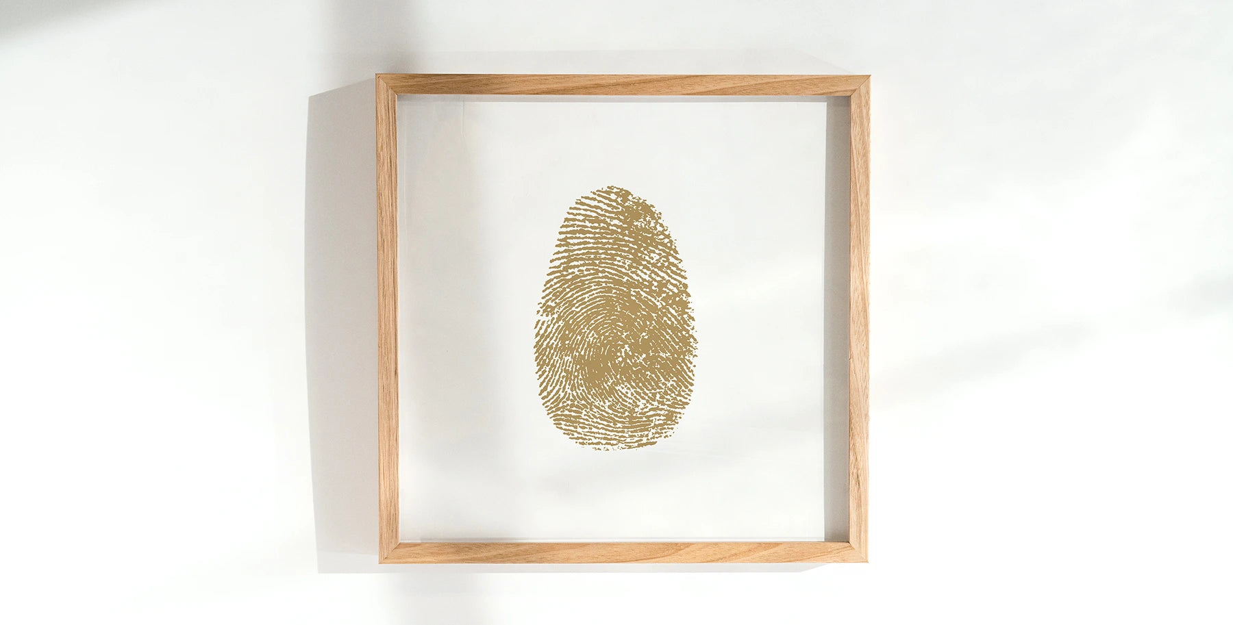 Framed artwork featuring a golden fingerprint centered on a white background. The light wood frame casts a soft shadow, creating a serene and minimalist feel.