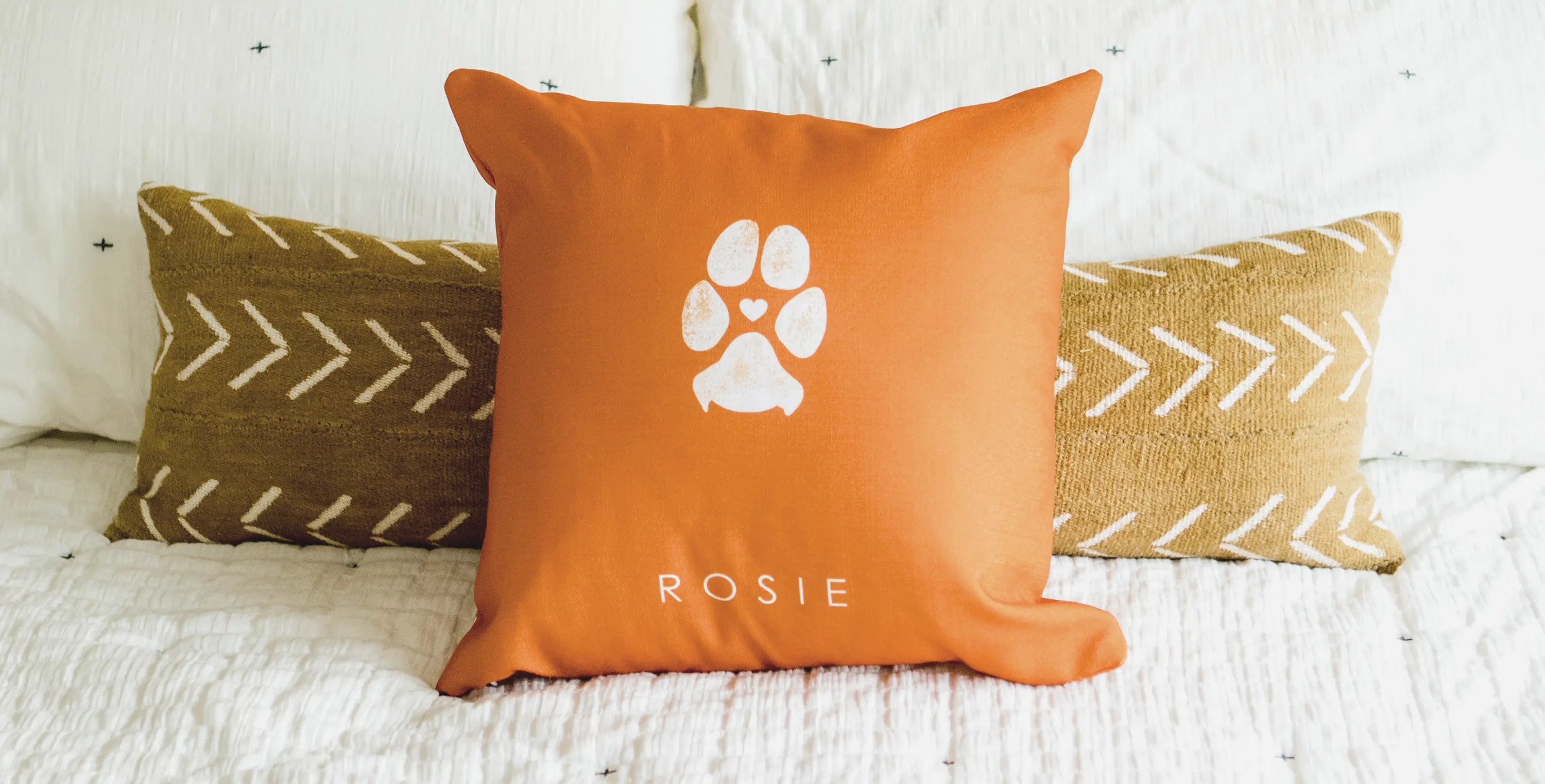 Orange cushion with a white paw print and heart, placed on a bed with two patterned brown pillows. The pet name 