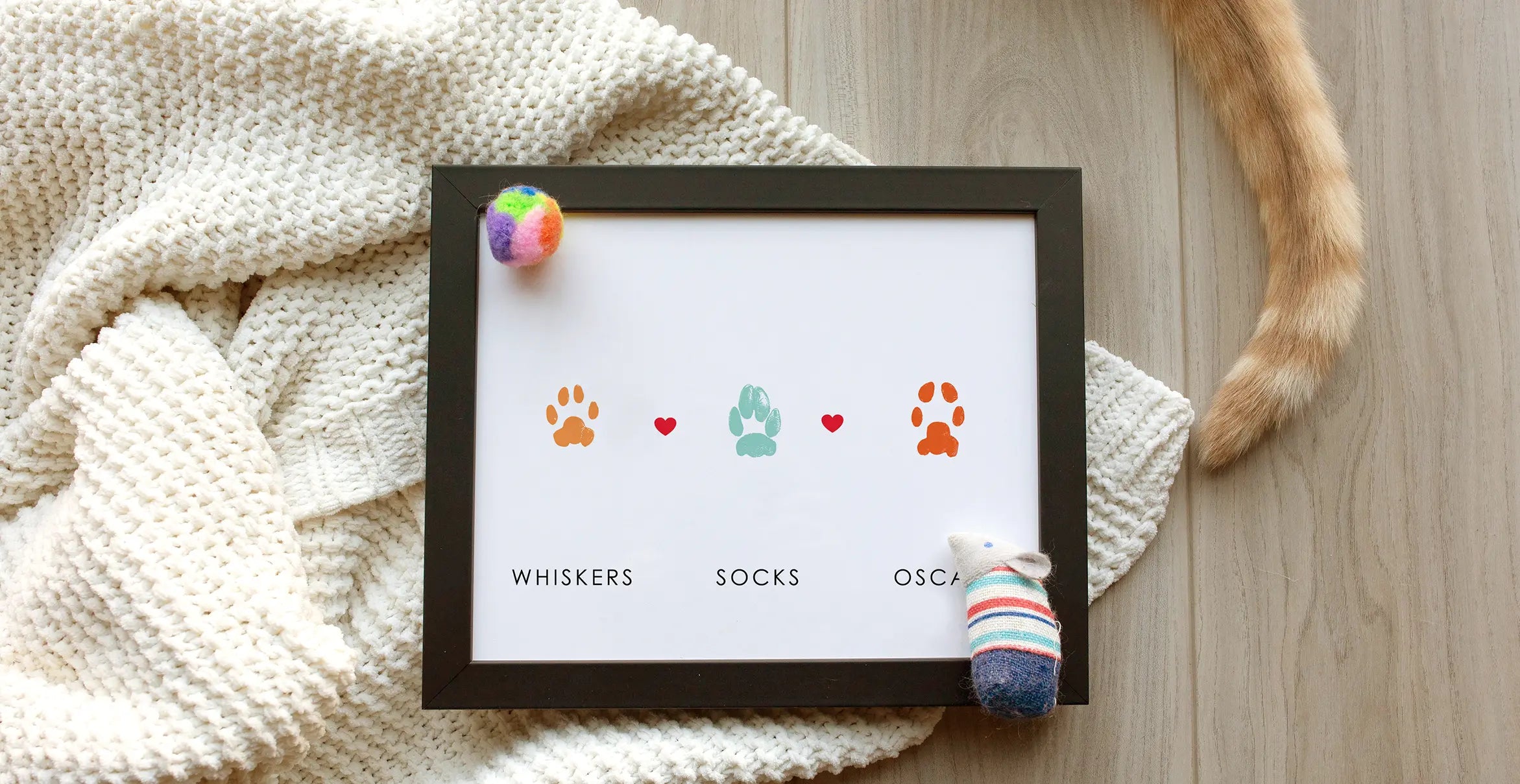 Framed print on wood floor shows three colorful cat paw prints with hearts above kitty names: Whiskers, Socks, Oscar. Cat tail, knit blanket, toys nearby. Cozy vibe.
