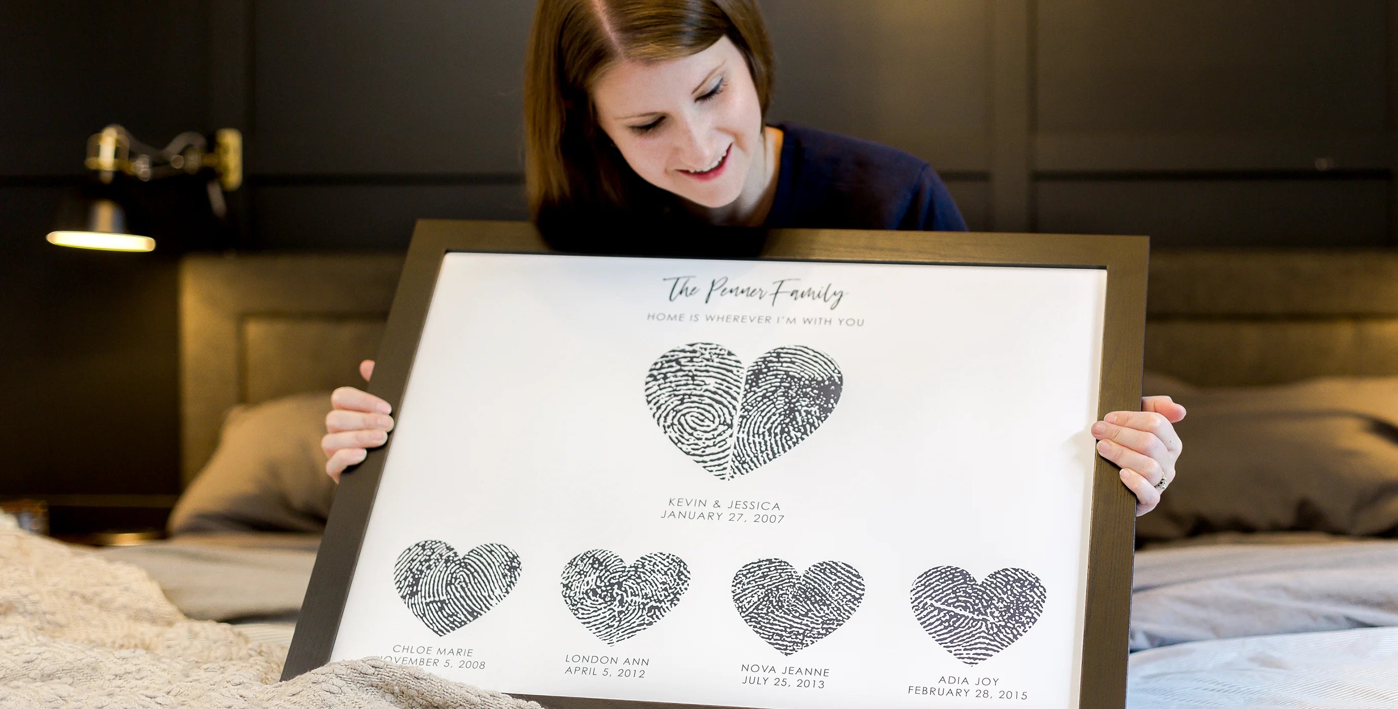 A woman sits on a bed, smiling while holding a framed piece of art featuring a family of fingerprint hearts. The room has a cozy and warm tone.