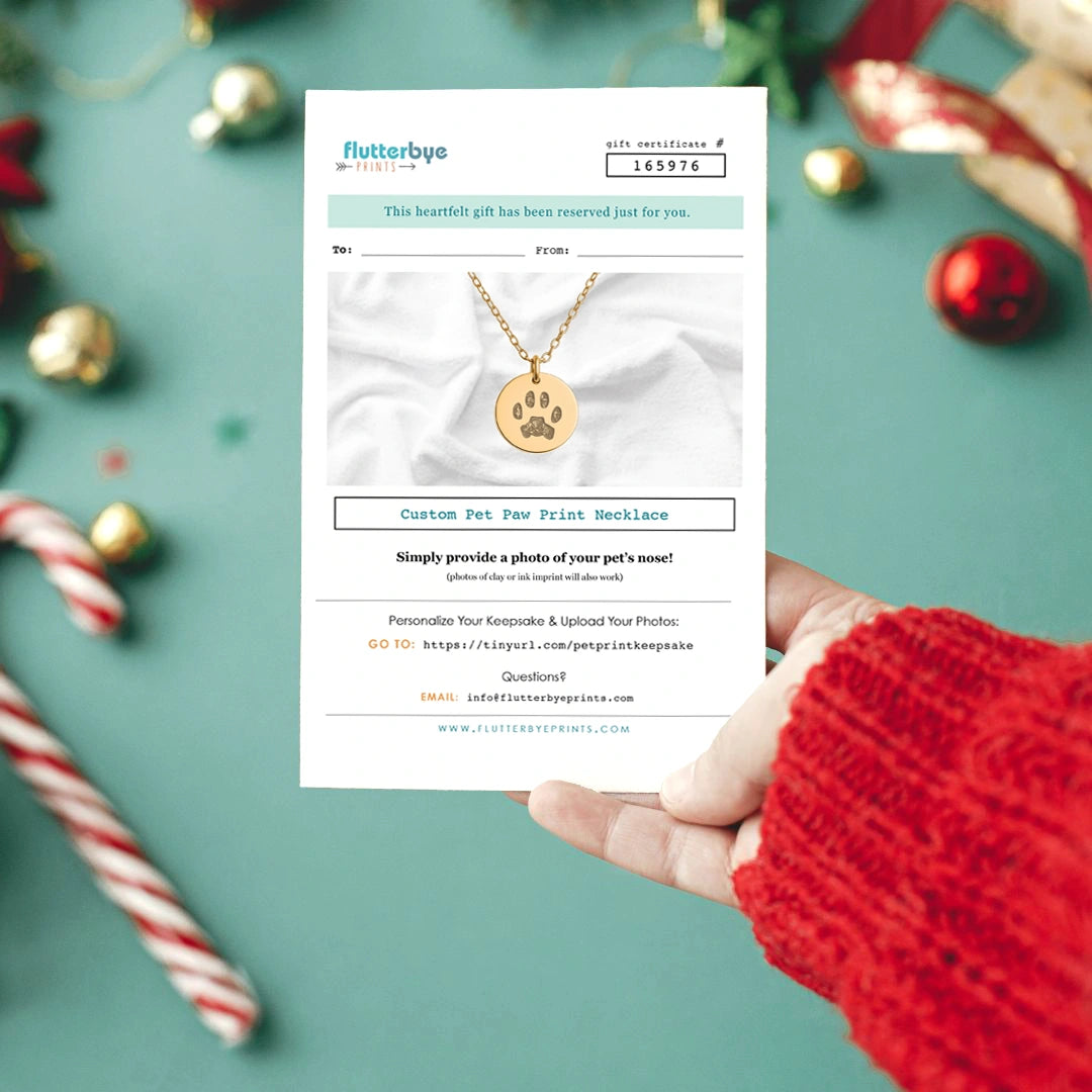 A hand holds a gift certificate for a custom pet dog nose print ornament, featuring a photo of dogs with a dogs nose print keepsake. Festive greenery adds a warm holiday feel.