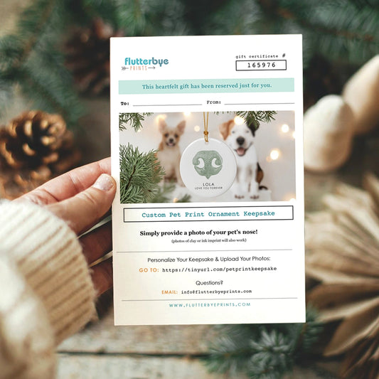 A hand holds a gift certificate for a custom pet dog nose print ornament, featuring a photo of dogs with a dogs nose print keepsake. Festive greenery adds a warm holiday feel.