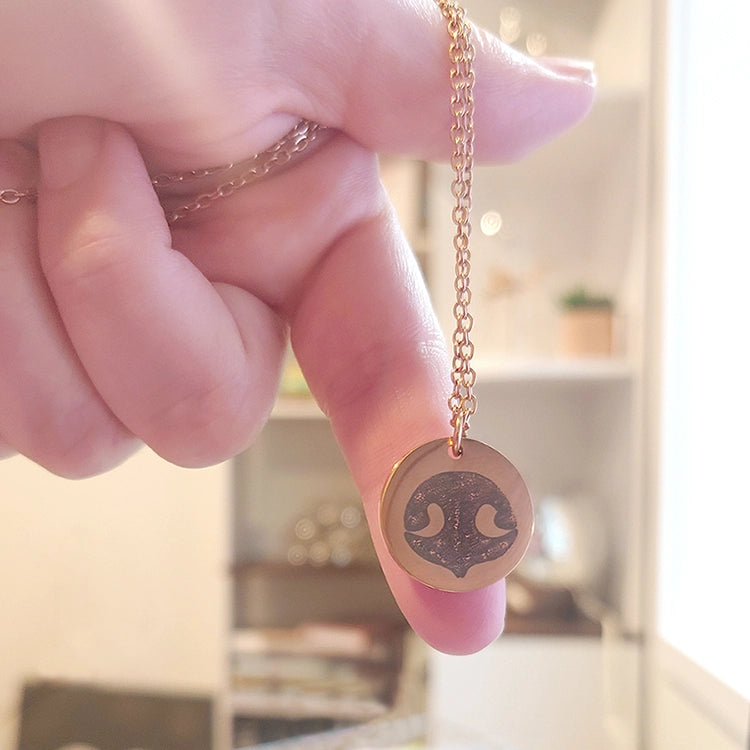 Hand holding a gold necklace with a circular pendant featuring a custom dog's nose print.
