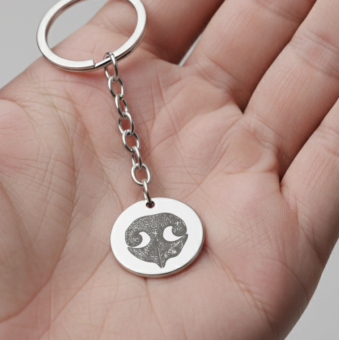A silver keychain with a round pendant featuring an intricately engraved design of dog nose print lies on an open palm. The focus is on the detailed metalwork.