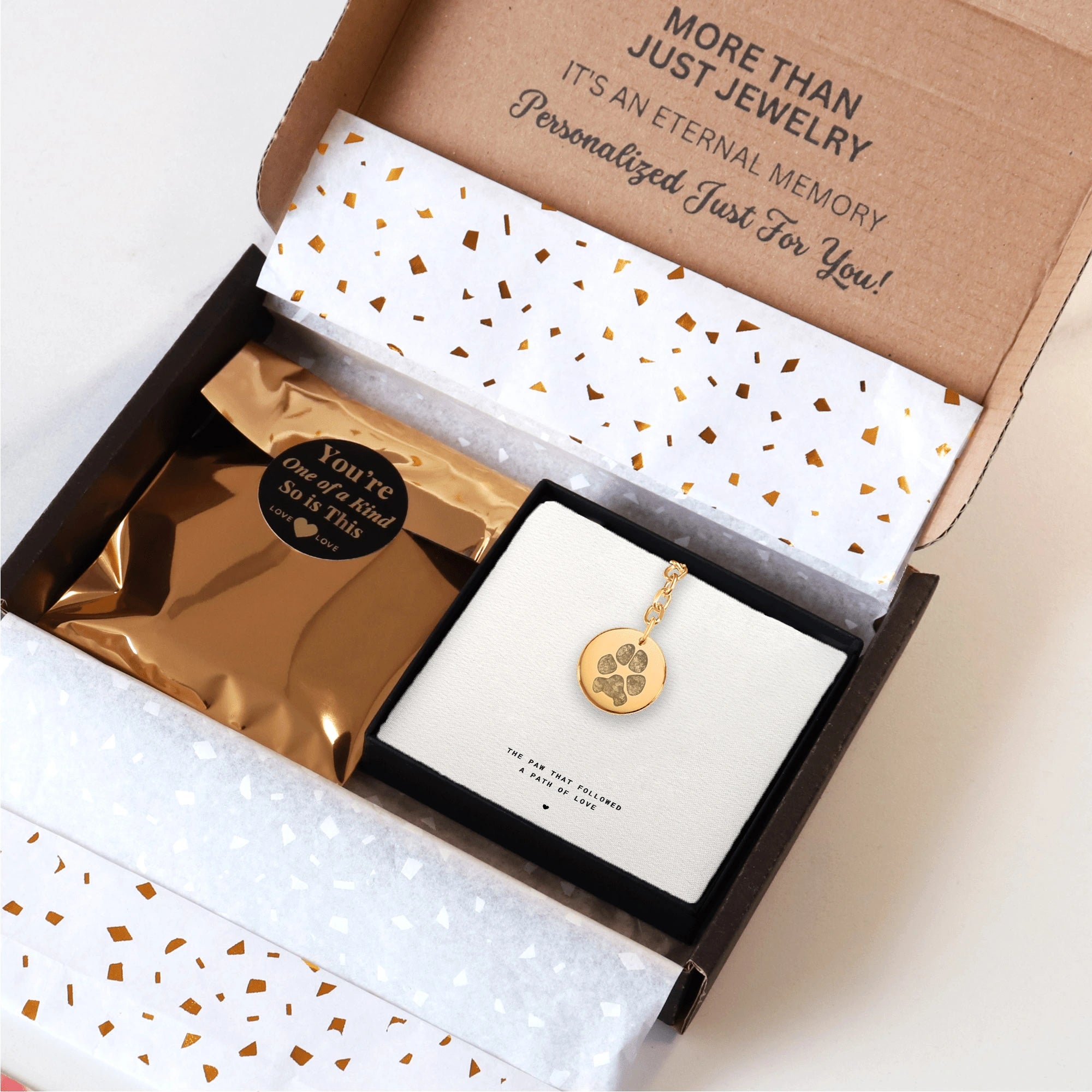 Open gift box with a gold keychain featuring a dog paw print pendant, surrounded by a glittery white and gold package.  Packaged and ready to give as a pet memorial gift.