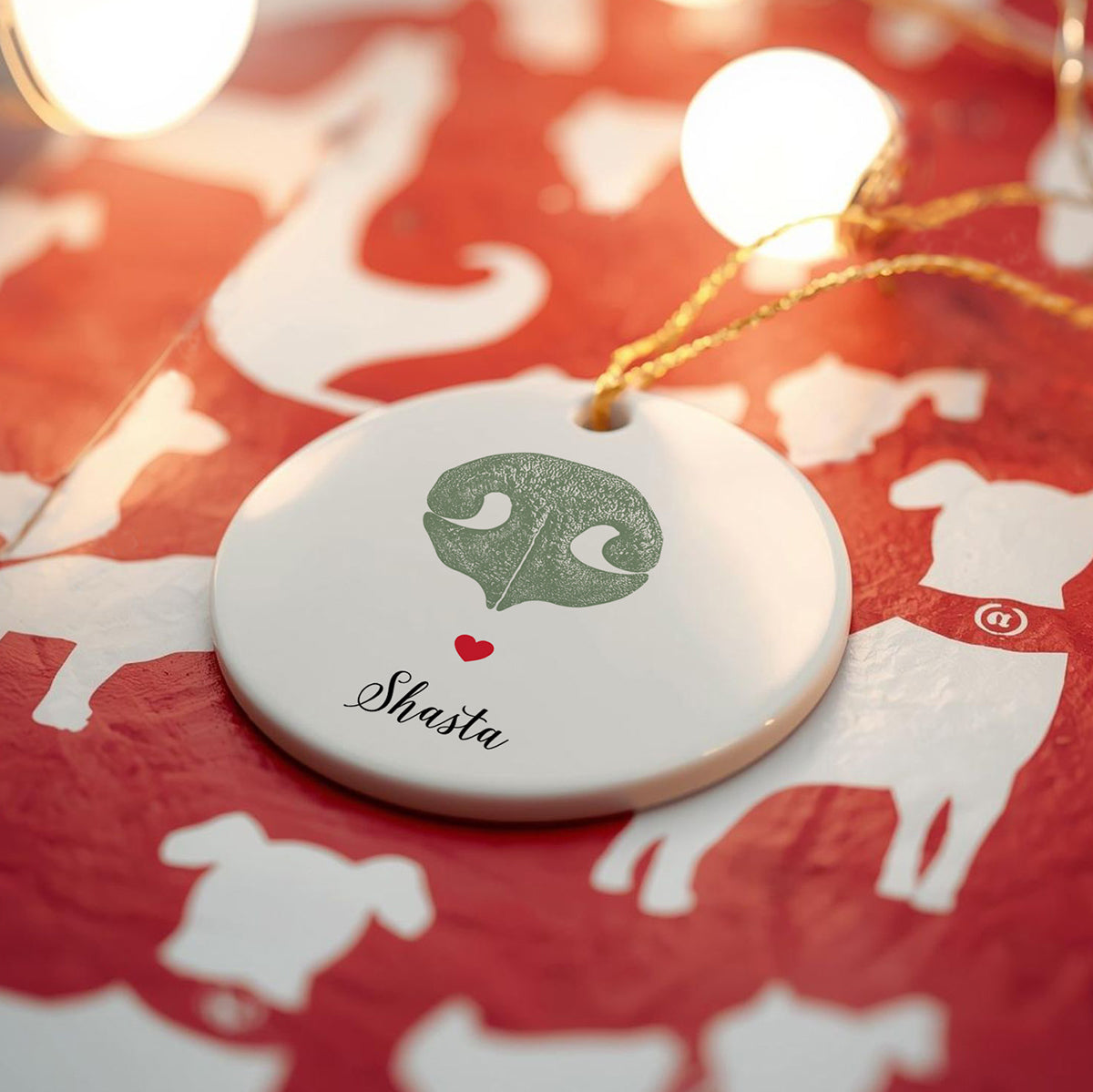 A round ceramic ornament with a green dogs nose print and the name "Shasta" written below, decorated with a small red heart. Soft lights illuminate the festive, red wrapping paper background adorned with white dog silhouettes. Cozy and warm holiday atmosphere.