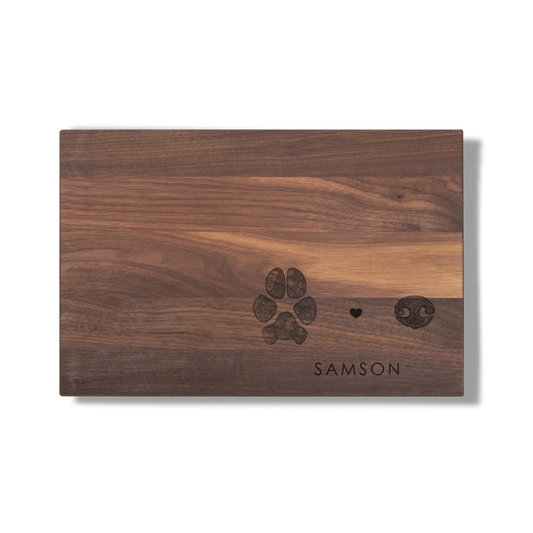 Wooden cutting board with a dark paw print, heart, and nose design engraved above the word "SAMSON." The board has a warm brown tone.