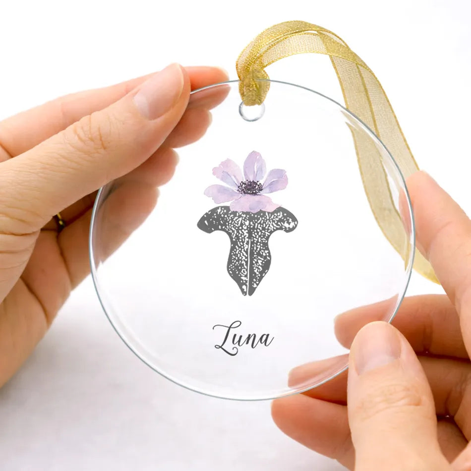 Two hands hold a round, clear ornament featuring a delicate purple flower and cat nose print. The word "Luna" is written below, with a gold ribbon attached.