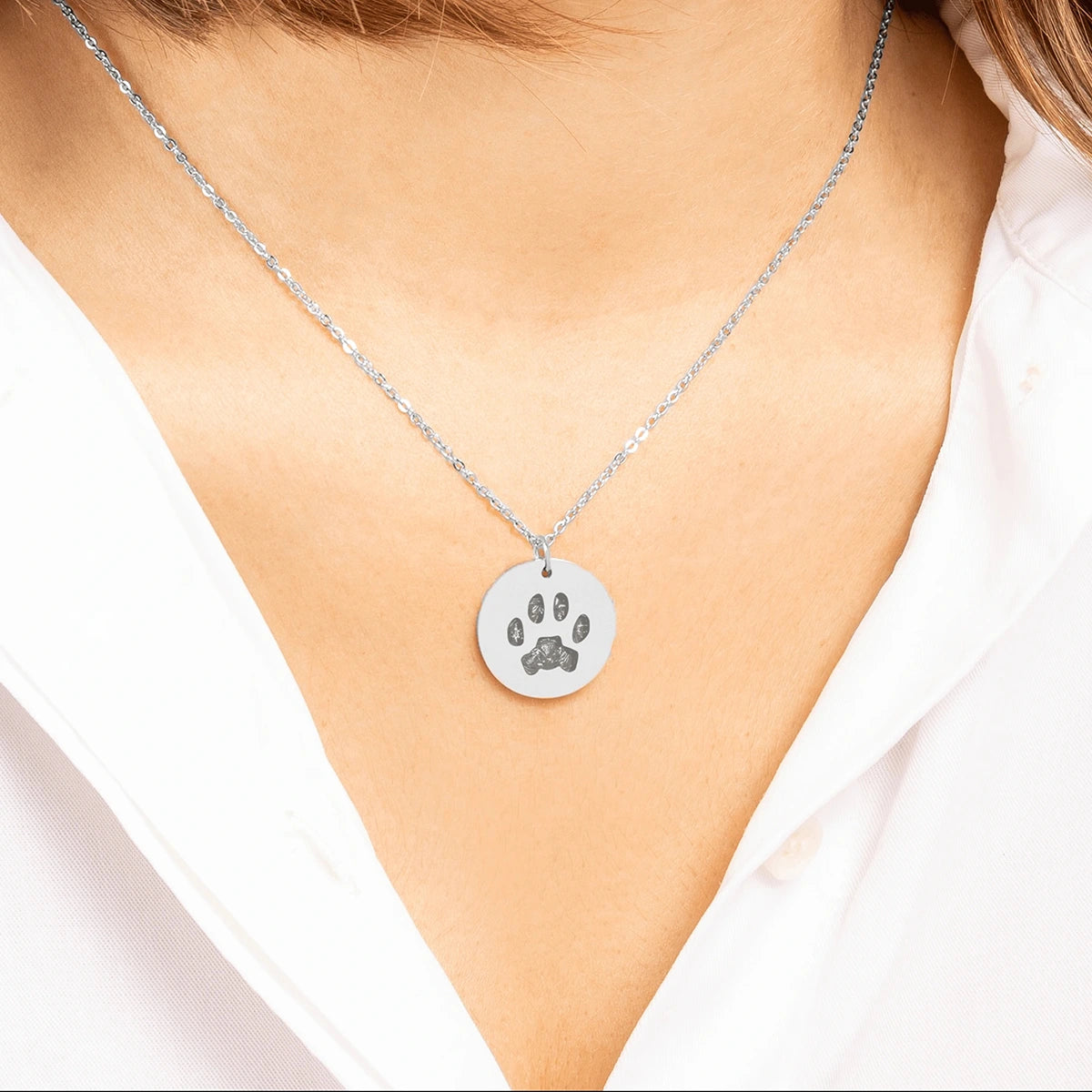 Close-up of a woman wearing a silver necklace with a round pendant featuring an engraved cats paw print pet memorial gift. She is dressed in a white shirt.