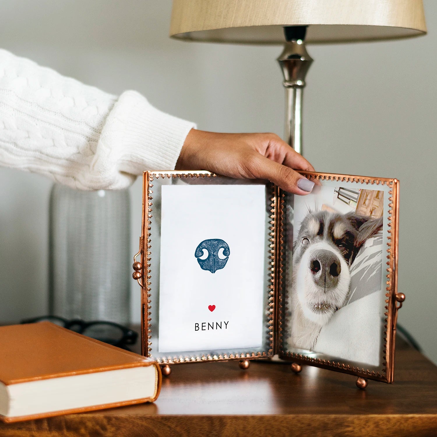 A hand holds a double photo frame on a wooden table. One side has a cute dog nose print design with the name "Benny"; the other shows a close-up of a dog's face and nose.