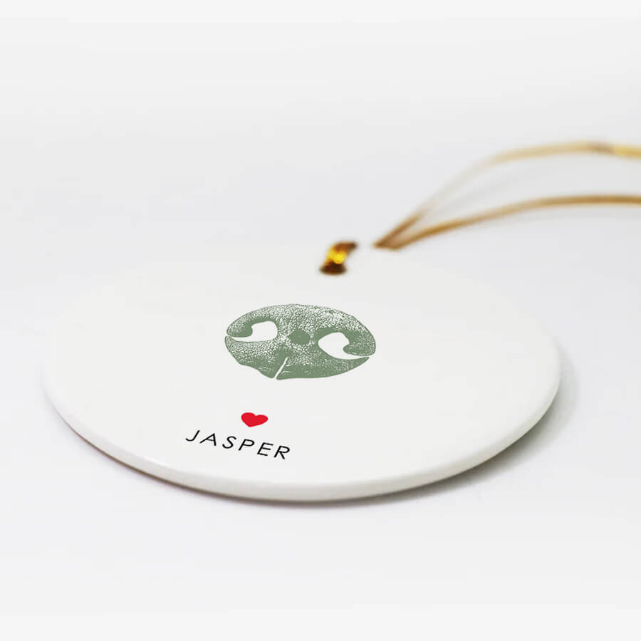 Round white ornament with a textured black dog nose print, small red heart, and the name “Jasper” beneath. Gold string for hanging attached at the top.