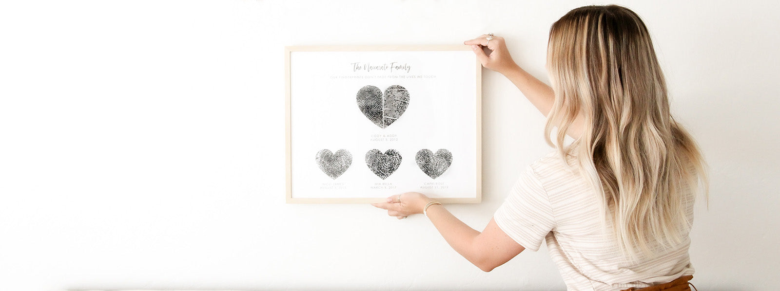 Woman hanging framed family fingerprint art on white wall.